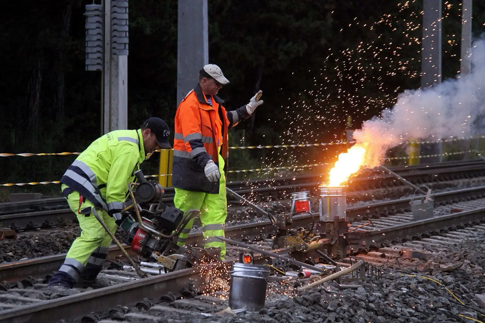 Thermite pour into a rail mould at dusk, crew in visors and PPE